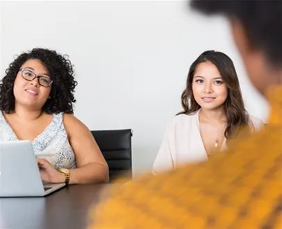 Two women sit at a desk smiling toward a third person who is mostly out of frame; this appears to be a professional setting, possibly a job interview.