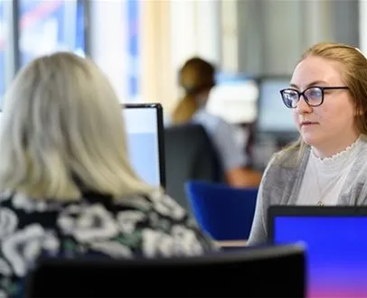 Two women working at computer desks in an office environment; the woman in focus wears glasses and listens attentively.